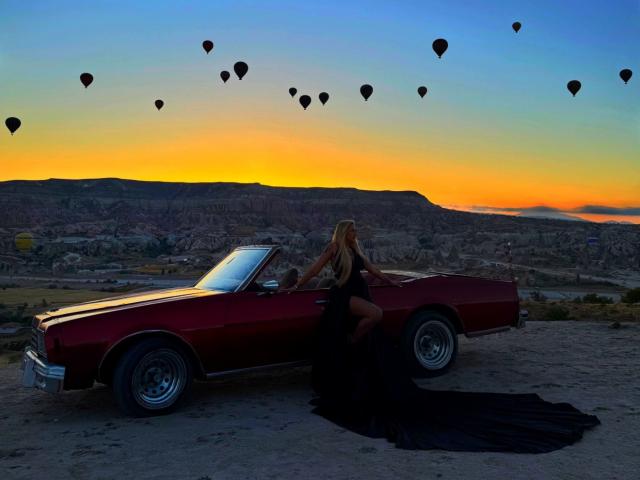 SinnersMuse with long blonde hair, black dress, and bare ass, leans on a red vintage car, surrounded by hot air balloons at sunset.