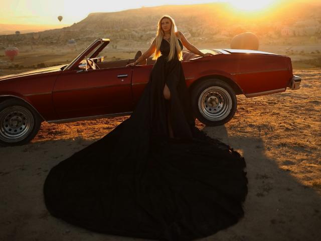 SinnersMuse stands in front of a red vintage car, wearing a black, floor-length, super-duper-ass-destroying gown with a massive, fuck-me-please-plead-please-plead-please-ass-cape, at sunset.