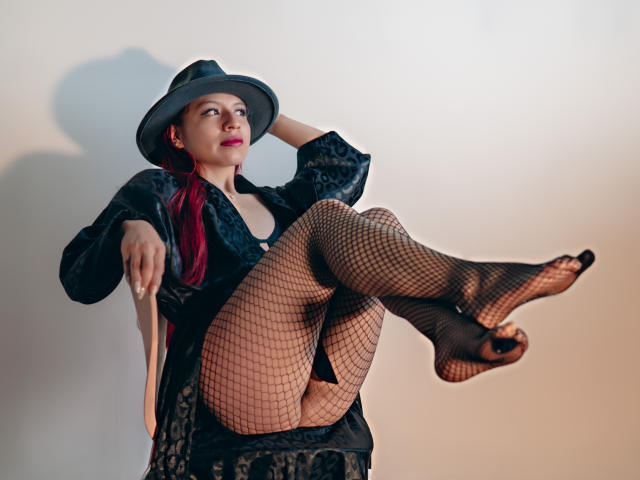 ZoeRedds with red hair, wearing a black hat, black lace dress, and fishnet stockings, sitting with legs up, exposing her black panties, and her tits slightly visible.