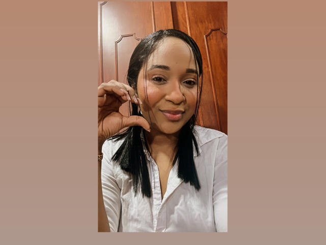 MelanyScoth with long black hair, white shirt, holding hair with her right hand, smiling, brown wooden background, soft light, gentle expression, slightly parted lips, casual look.