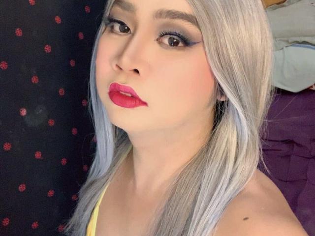 AsianFemboyX has pale skin, long silver hair, dark eyeliner, pink lipstick, and a yellow top, showing a cute, confident face with slightly parted pink lips.