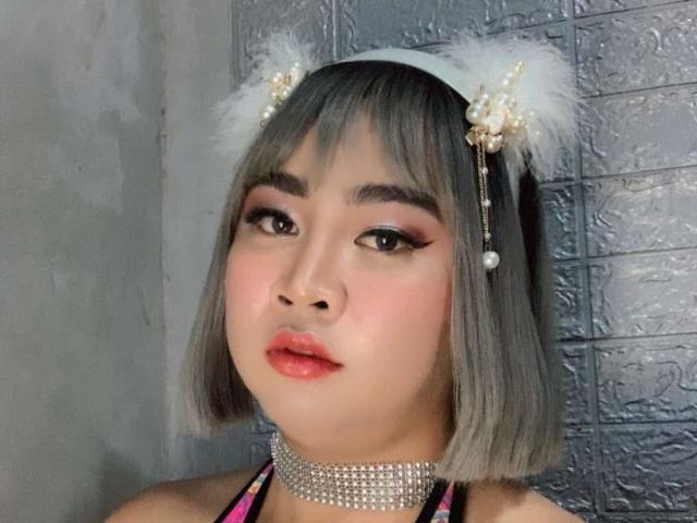 AsianFemboyX with gray bob, white fluffy cat ears, pearl choker, pink lipstick, and heavy eyeliner, looking at camera. Silver-tiled wall in background.