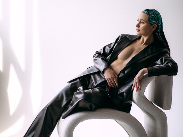 AlexaWilds in a black leather outfit, sitting on a white chair, revealing her bare boobs, with teal braids, red nails, and a confident, sexy look.