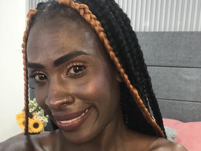 SamaraaJackson69 has dark skin, black braided hair with orange tips, a nose ring, and is smiling with glossy eyes, showing her bare shoulders and a pink pillow.