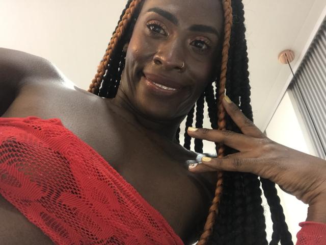 SamaraaJackson69 with dark skin, orange and black braided hair, smiling, wearing a red lace top, showing her tits, nose ring, and touching her face with green nails.