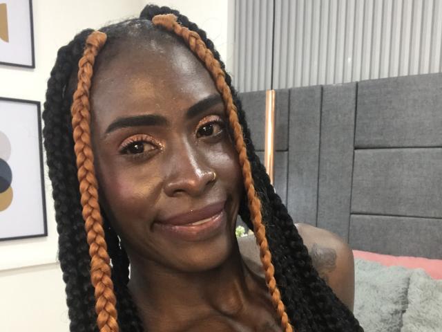 SamaraaJackson69 mit dunkler Haut, orange-schwarzen Zöpfen, kleiner Nase, lächelnd, glänzendes Gesicht, braune Augen, dezentes Tattoo an der rechten Schulter, graue Kopfteil im Hintergrund..