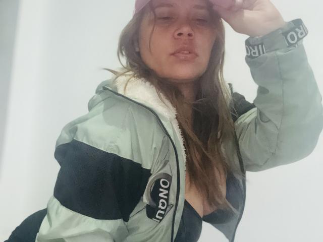 SophiaQueens trägt eine grüne und schwarze Jacke, eine rosa Mütze und ein schwarzes Oberteil. Sie hat ihre Hand auf dem Kopf und zeigt dabei leicht ihre Brüste. Sie hat braunes Haar und einen neutralen Gesichtsausdruck..