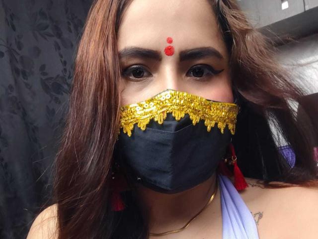DaliaDubey has brown wavy hair, a red forehead dot, black mask with gold trim, red tassels, and a white halter top, showing a small tattoo on her shoulder.