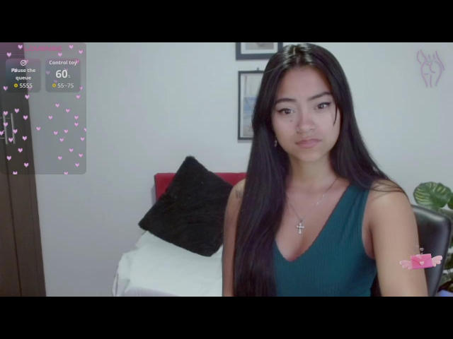NatalyEscobar hat langes, schwarzes Haar, trägt ein grünes Top ohne Ärmel, das ihre kleinen Brüste zeigt, und eine Kreuzkette. Sie sitzt auf einem schwarzen Kissen und einer weißen Decke..