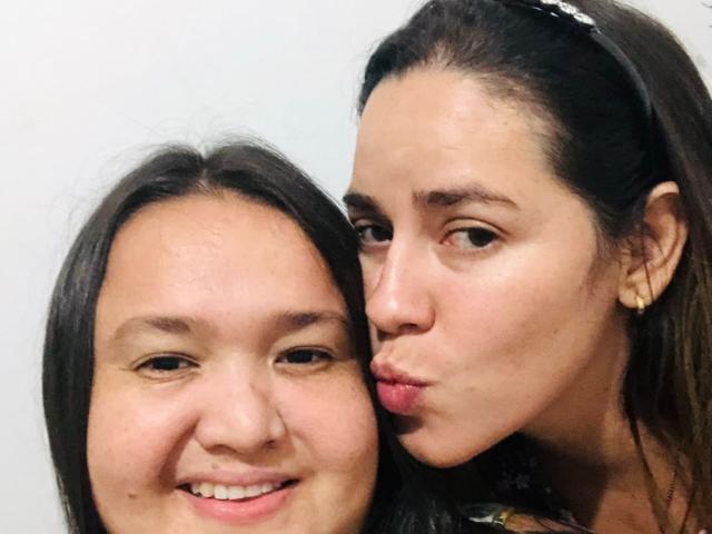 CarlaNaughty with puffy ass kissing CarlaNaughty's cheek, CarlaNaughty on the left smiling, CarlaNaughty on the right puckering lips, both have dark hair, CarlaNaughty's face is closer.