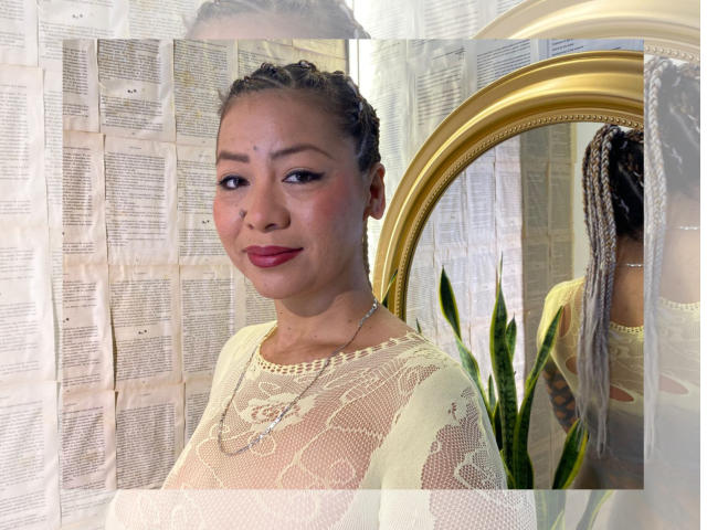 MirandhaX with braided hair, light skin, pink lipstick, wearing a sheer white lace top, gold necklace, and a gold mirror in the background. Her tits are partially visible.