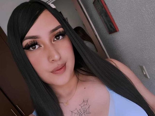 Chloey69 with long black hair, full eyebrows, big eyelashes, light skin, nose piercing, lip piercing, blue top, spider web tattoo on chest, smiling.