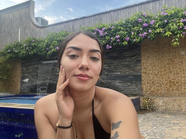 SharonKlart, com cabelo castanho, top de alça preta, mão no rosto, tatuagem de borboleta, piscina azul ao fundo, flores roxas, parede de pedra, sorriso discreto, pele morena, pulseira preta..