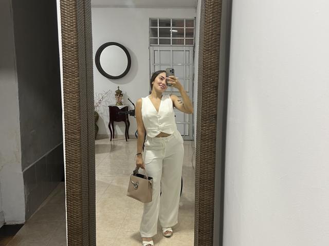 SharonKlart, white outfit, sleeveless top, high-waisted pants, holding tan bag, standing in front of mirror, smiling, brown hair, white open-toe heels, reflection, attractive, sexy.
