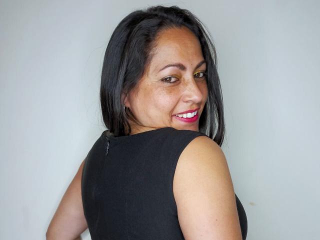 ScarlettBarness with black hair, brown eyes, pink lipstick, and black sleeveless top, looking back over her shoulder, smiling. Her medium-sized tits are partially visible from the side.