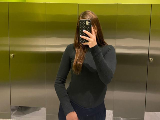 EvaDaMore portant un haut noir à manches longues, tenant un téléphone avec une coque noire à pois, cachant son visage, avec de longs cheveux bruns, dans une cabine de toilettes..