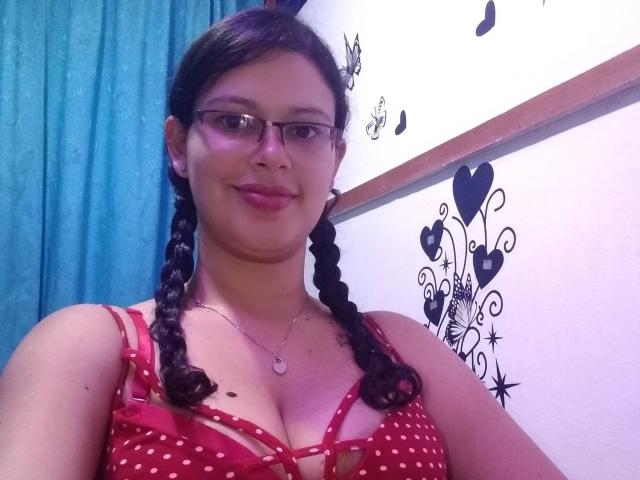 KimValentina, belle avec des lunettes, cheveux noirs tressés, haut rouge à pois, dévoilant sa poitrine, souriante, rideau bleu, autocollants muraux noirs, collier en argent avec pendentif en forme de cœur..