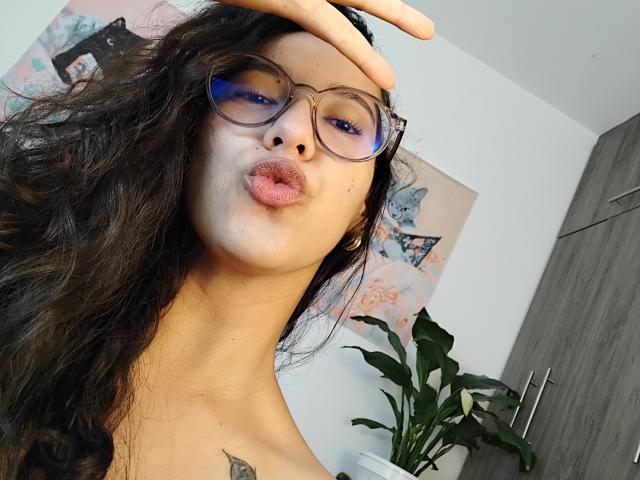 Mllevv has pouty lips, blue-tinted glasses, and a hand on her head, with wavy black hair, a tattoo on her chest, and a plant in the background.
