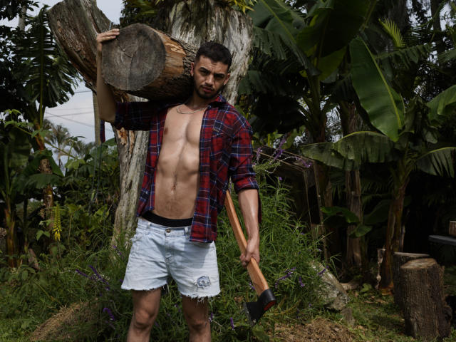 MichaelMagno, shirtless with a red plaid flannel, holds a log over his shoulder and a axe in his hand, wearing white denim shorts, standing in a forest.
