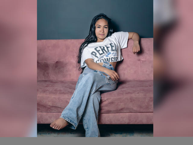 AddaCortez with long black hair, white "Perfect" t-shirt, and blue jeans, leaning on pink sofa, barefoot, right leg crossed over left, relaxed, confident, cute, casual.