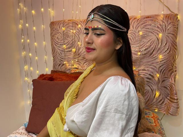 NayaYalitza with dark hair, yellow dress, white shawl, and jeweled headpiece, showing her tits, pink lips, and bold eyeliner, smiling, surrounded by warm fairy lights.