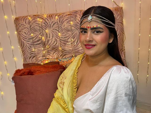 NayaYalitza with medium tits, wearing a yellow sari and white blouse, has dark hair, pink lipstick, and a jeweled headpiece, smiling, with fairy lights in the background.
