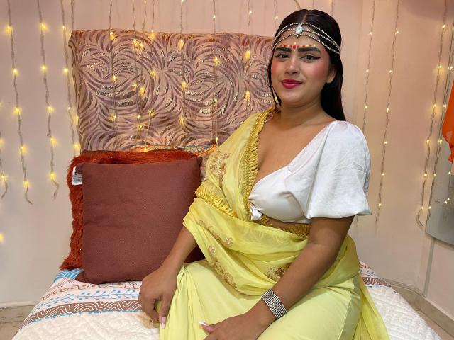NayaYalitza with medium tits, wearing a yellow sari, white blouse, and gold jewelry, sitting on a bed with red pillows, string lights, and a patterned wall.