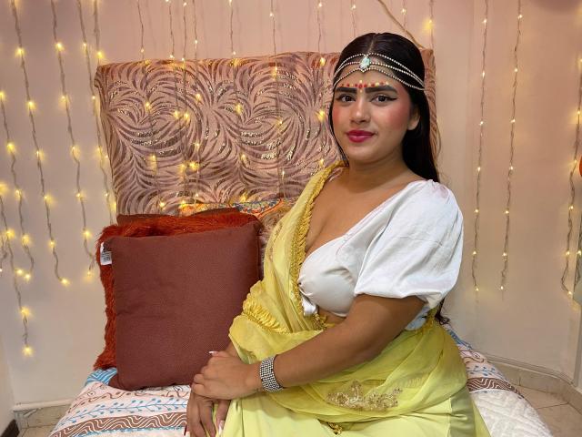 NayaYalitza, a curvy woman with medium brown skin and dark hair, wears a yellow saree, white blouse, and jeweled headpiece, sitting on a bed with a red pillow.