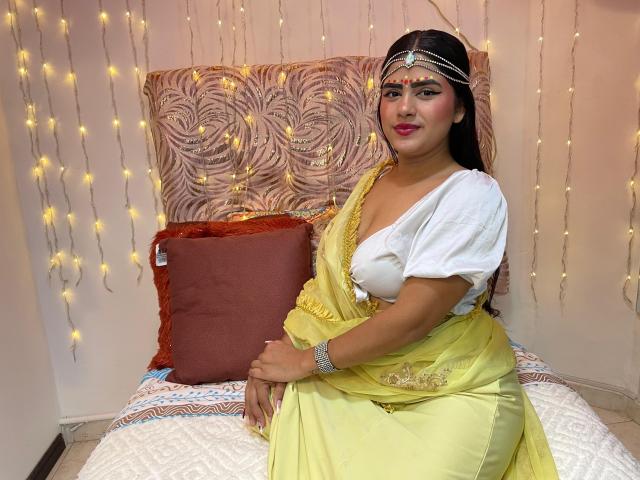 NayaYalitza with dark hair, wearing a yellow sari, white top, and headpiece, sitting on a bed with a red pillow, hands on her ass, fairy lights in the background.