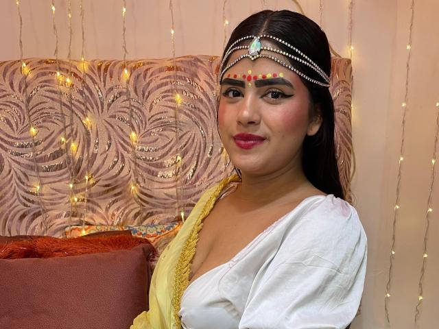 NayaYalitza with dark hair, wearing a white top and yellow dress, has a jeweled headpiece, pink lipstick, and a subtle smile, showing her cute tits and pretty ass.