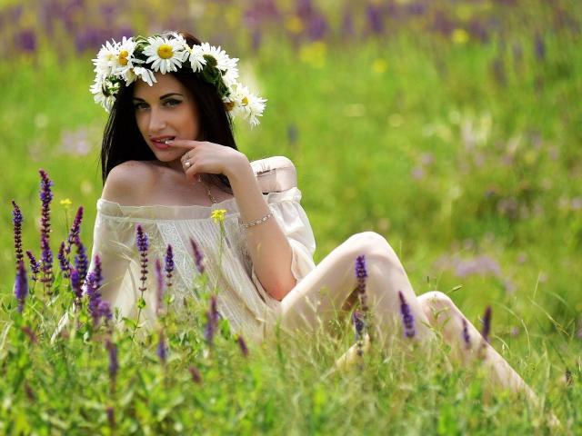 MiaDefne with white daisy headpiece, off-shoulder cream dress, sitting in green field, touching her chin, purple and yellow flowers around, smiling softly, long dark hair, green eyes.