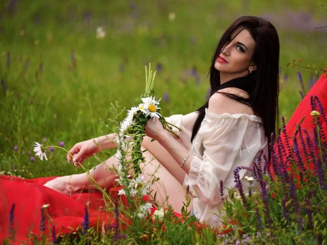 MiaDefne with long dark hair, white off-shoulder top, holding white flowers, sitting on red cloth, green grass, purple and white flowers, looking back with red lips.