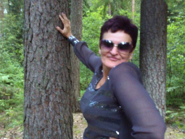 PaulaGoldens with short black hair, wearing dark sunglasses and a sheer gray top, holding a tree with her right hand, smiling at the camera, showing her tits slightly.