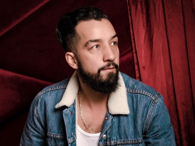 Decimo69, con una barba espesa, cabello oscuro y una chaqueta vaquera con un cuello de piel blanca, vistiendo una camiseta blanca y un collar de cadena, mirando hacia un lado..