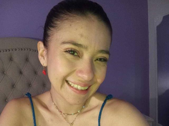 CattsyCathy has a smile with slightly crooked teeth, wearing a blue strap top, and a pink earring. She has a light skin tone and brown hair in a bun.
