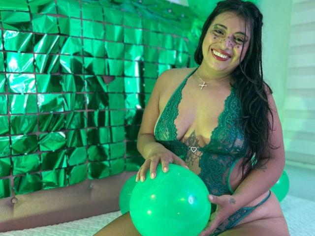 TammyyBrown with green lace lingerie, holding a green balloon, smiling, has tattoos on her chest and side, long black hair, wearing a cross necklace, green room lighting.