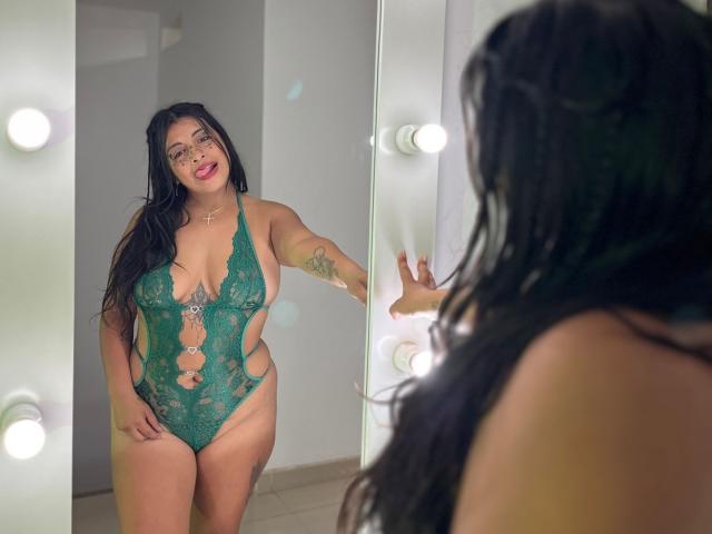 TammyyBrown with long black hair, green lace lingerie, showing her big tits and curvy ass, looking in a mirror with lights, tattoo on her right arm, smiling.