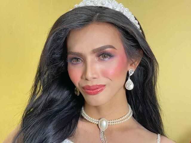 KarenCollins has dark, wavy hair, pink blush, and red lipstick, wearing a pearl necklace, earrings, and a diamond tiara, with purple eyeshadow and a slight smile.