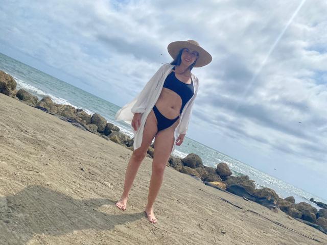 EmilyBonnett debout sur une plage rocheuse, vêtue d'une combinaison noire, laissant apparaître sa coupe de cheveux courte, avec une chemise blanche ouverte et un chapeau de soleil, souriant à l'appareil photo..