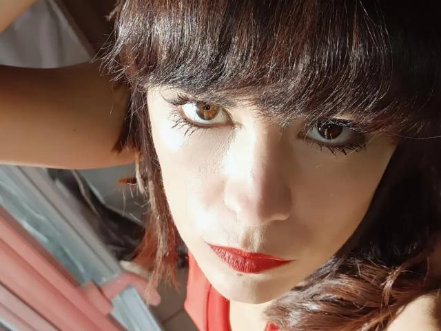 OrianaLaFrancaise with dark brown eyes, red lipstick, and medium-length brown hair, looking up with a serious expression, wearing a red top. Her eyelashes are long and curled.