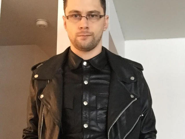 FetishCuir69, wearing black leather jacket with silver zippers, black leather shirt, and glasses, has short dark hair, light skin, and a slight beard, standing in a white room.