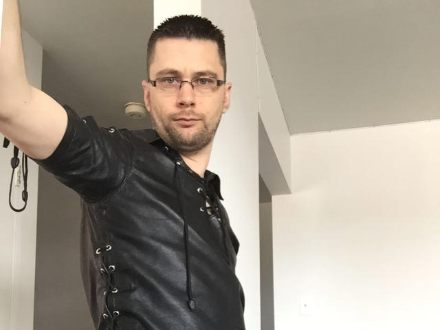 FetishCuir69, short brown hair, glasses, black leather shirt with lace-up sides, slight beard, raising arm, white wall, ceiling light in background.