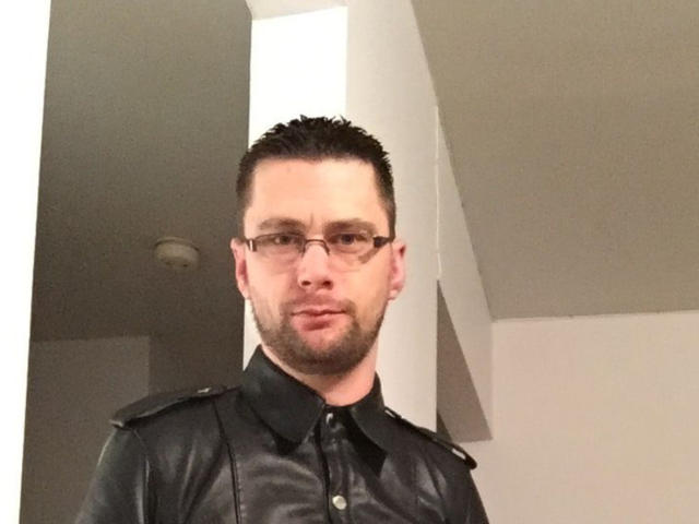 FetishCuir69, a nerdy dude with glasses, short dark hair, and a beard, wearing a black leather shirt, looking serious. Boobs: no, ass: no, pussy: no, tits: no.