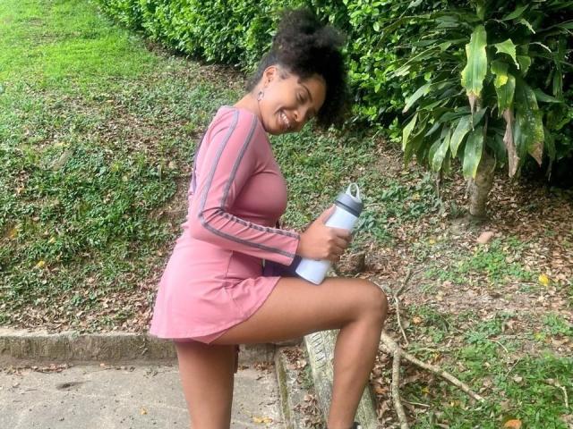 MeganWlliams with dark skin, curly hair, wearing a pink dress, holding a water bottle, kneeling with one leg up, smiling, showing her tits, in a green, leafy area.