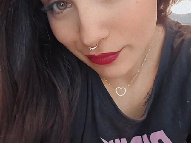 AshleyDaphna met rode lippenstift, een neusring, een hartvormige ketting, een tatoeage aan de rechterkant, een zwart shirt, bruin haar en een lichte glimlach, waardoor haar schattelijke, aantrekkelijke gezicht te zien is..