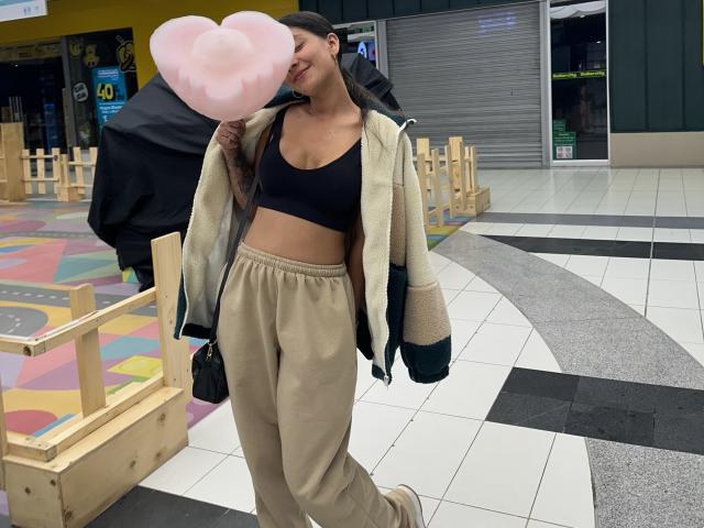 ThaliaAres with pink heart-shaped puff candy, black sports bra, beige jacket, tan pants, black bag, smiling, black cap, holding candy near her mouth, standing in a tiled hallway.