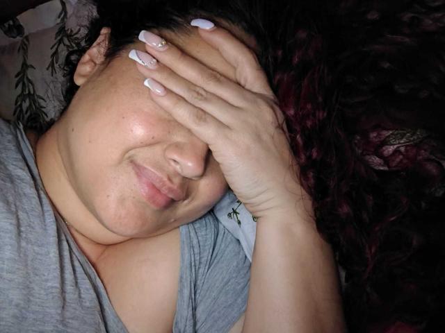 Le visage de Lalitzfet est partiellement dissimulé par sa main, laissant apparaître un léger sourire. Elle a une peau pâle, des cheveux noirs et bouclés, et un vernis à ongles blanc. Son t-shirt gris révèle ses seins..