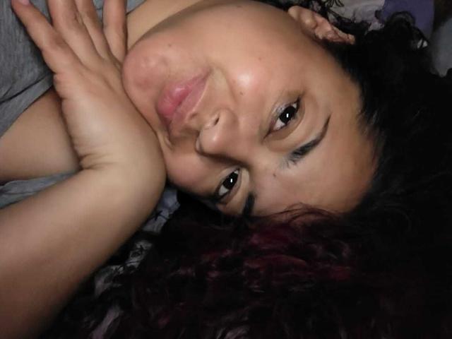 Lalitzfet, met volle, roze lippen, bruine ogen en donker krullend haar, leunde met haar hoofd op haar hand, droeg een grijze trui en keek op met een subtiele glimlach..
