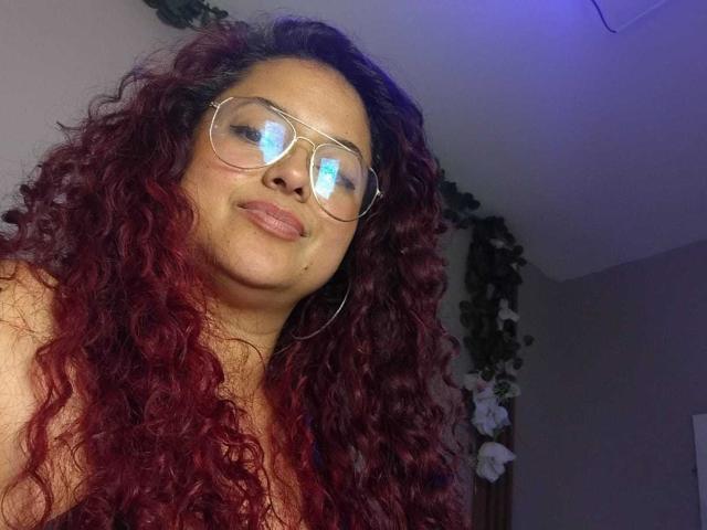 Lalitzfet, avec ses cheveux roux bouclés, portant des lunettes, souriant légèrement, montrant ses seins dans un selfie. Son reflet se voit dans ses lunettes. Elle est à l'intérieur, avec une lumière violette douce..
