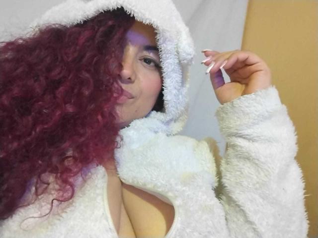 Lalitzfet met krullend rood haar, een witte, pluizige hoodie, en opvallende grote borsten, met make-up, die haar vinger tegen haar wang houdt en glimlacht..